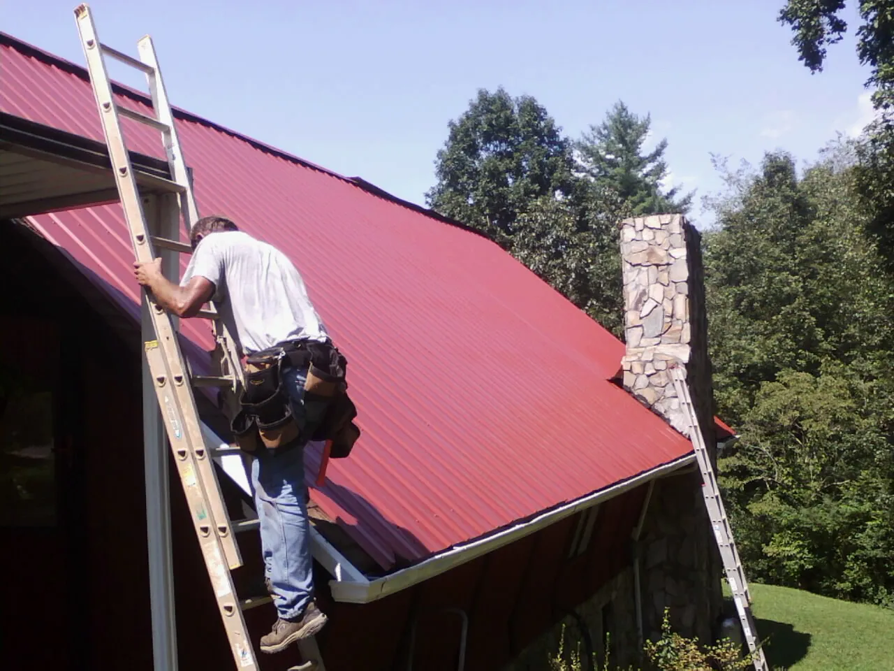 Roofer on a ladder beside a red metal roof and stone chimney — Soffit & Fascia Repair by Roofing Brothers in Lake Junaluska
